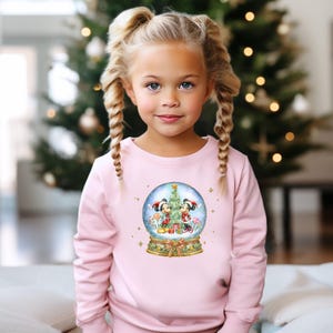 May include: A pale pink sweatshirt with a snow globe design featuring Mickey and Minnie Mouse in festive Christmas outfits. The snow globe design includes a Christmas tree, presents, and candy canes. The sweatshirt is worn by a young girl with blonde plaits.
