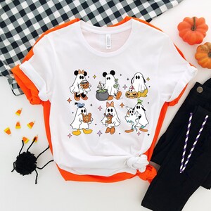 May include: White t-shirt featuring a graphic of Disney characters dressed as ghosts for Halloween. The characters include Mickey Mouse, Minnie Mouse, Donald Duck, Goofy, and Pluto.