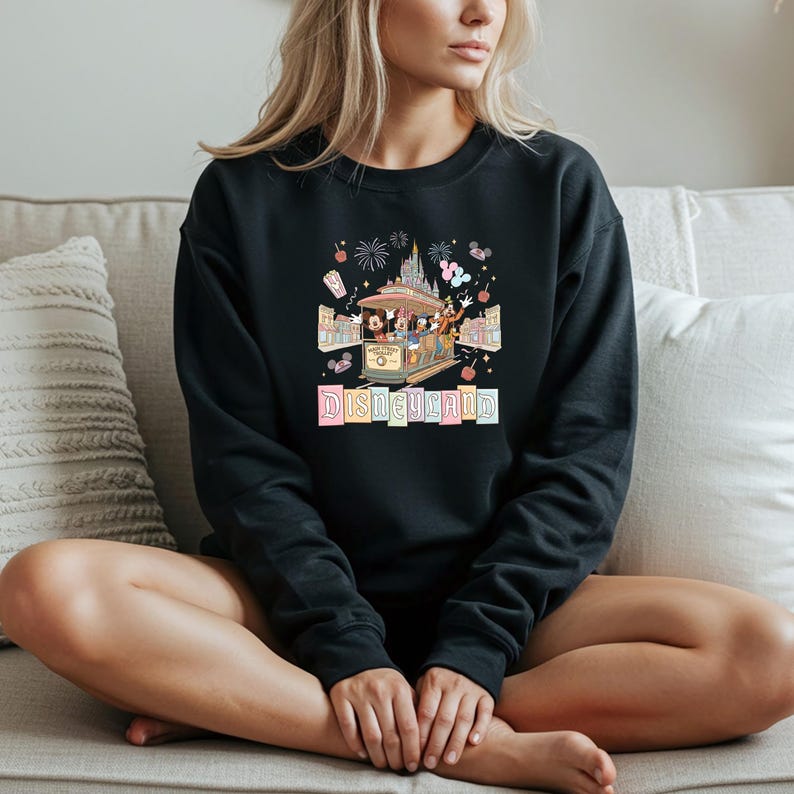May include: Black sweatshirt featuring a colorful Disneyland graphic. The graphic includes a trolley with Disney characters, fireworks, and the word "Disneyland" in pastel letters. The sweatshirt is a crew neck style.