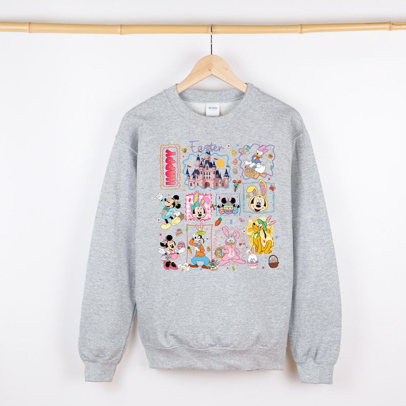 May include: A light gray sweatshirt featuring a colorful Easter-themed graphic. The design includes various Disney characters, a castle, and the words "Happy Easter". The sweatshirt is hanging on a wooden hanger.