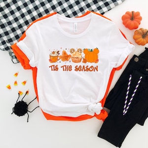 Pu&ograve; includere: T-shirt bianca con bordi arancioni con una grafica a tema autunnale. La grafica include un latte alla zucca, una foglia d'acero, un cupcake e una zucca. Il testo "Tis The Season" &egrave; stampato in arancione.