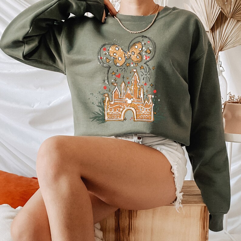 May include: A green sweatshirt with a gingerbread castle and a bow graphic. The castle is decorated with red and white icing and has a Mickey Mouse head on the front. The bow is red and white with gold glitter.