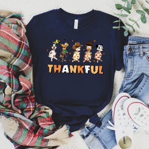May include: Navy blue t-shirt featuring a graphic of cartoon dogs in festive attire, with the word "THANKFUL" in orange and white. The dogs are dressed in various outfits, including cowboy hats and fall-themed patterns. A casual, seasonal design.