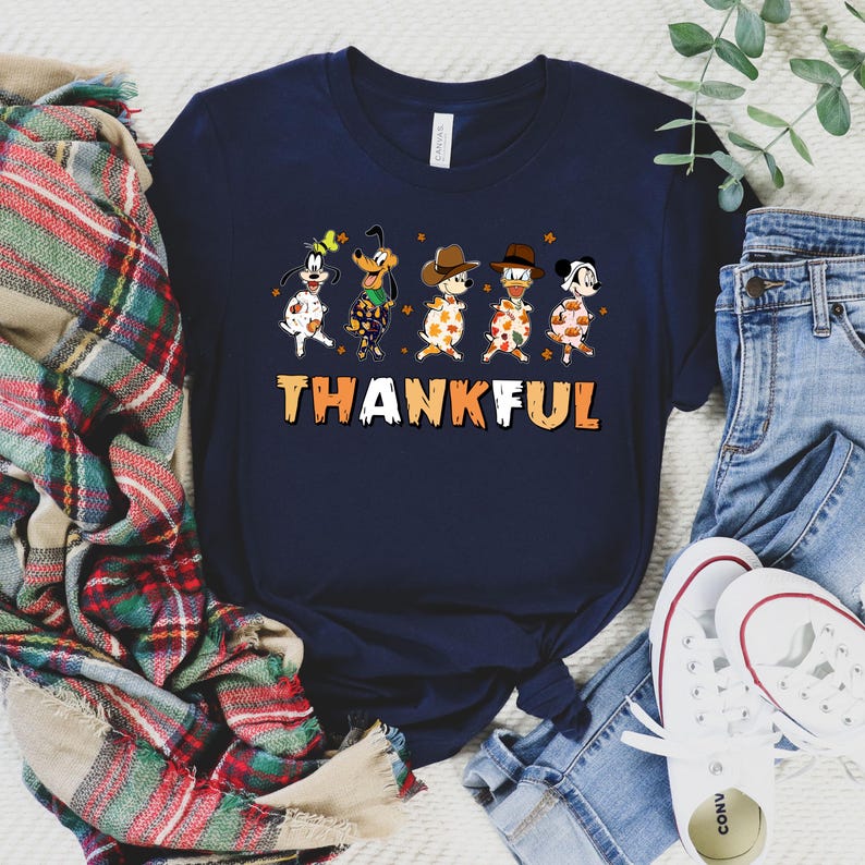 May include: Navy blue t-shirt featuring a graphic of cartoon dogs in festive attire, with the word "THANKFUL" in orange and white. The dogs are dressed in various outfits, including cowboy hats and fall-themed patterns. A casual, seasonal design.