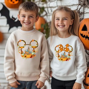 Może przedstawiać: Dwoje dzieci w kremowych i białych bluzach z motywami Halloween. Jedna bluza ma głowę Myszki Miki z zamkiem, dyniami i jesiennymi liśćmi. Druga ma kokardę Myszki Minnie z zamkiem i dyniami.
