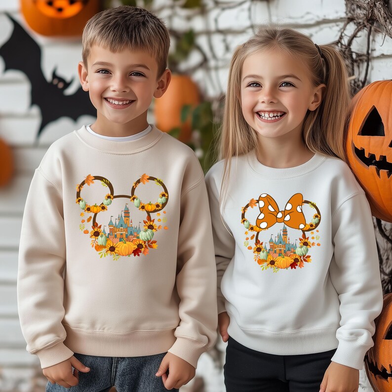 Może przedstawiać: Dwoje dzieci w kremowych i białych bluzach z motywami Halloween. Jedna bluza ma głowę Myszki Miki z zamkiem, dyniami i jesiennymi liśćmi. Druga ma kokardę Myszki Minnie z zamkiem i dyniami.