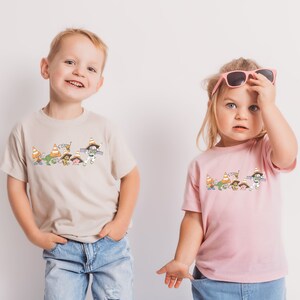 Op de afbeelding: Twee kinderen dragen T-shirts met een cartoonafbeelding van figuren die verkeerskegels dragen. De jongen draagt een beige T-shirt en het meisje draagt een roze T-shirt.