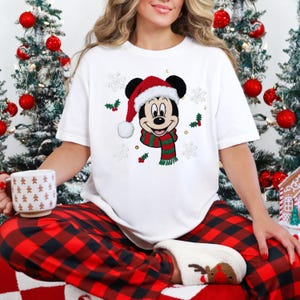 May include: White t-shirt featuring a festive Mickey Mouse design. Mickey is wearing a Santa hat and a green and red striped scarf. The shirt is decorated with snowflakes and holly. The person is wearing red and black plaid pajama pants.