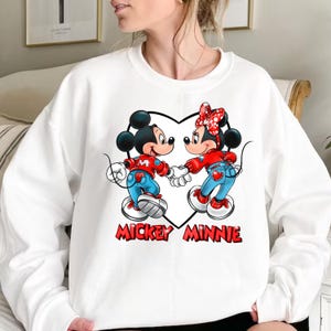 May include: White sweatshirt with a graphic of Mickey Mouse and Minnie Mouse holding hands inside a heart. The text "Mickey Minnie" is printed below the graphic.