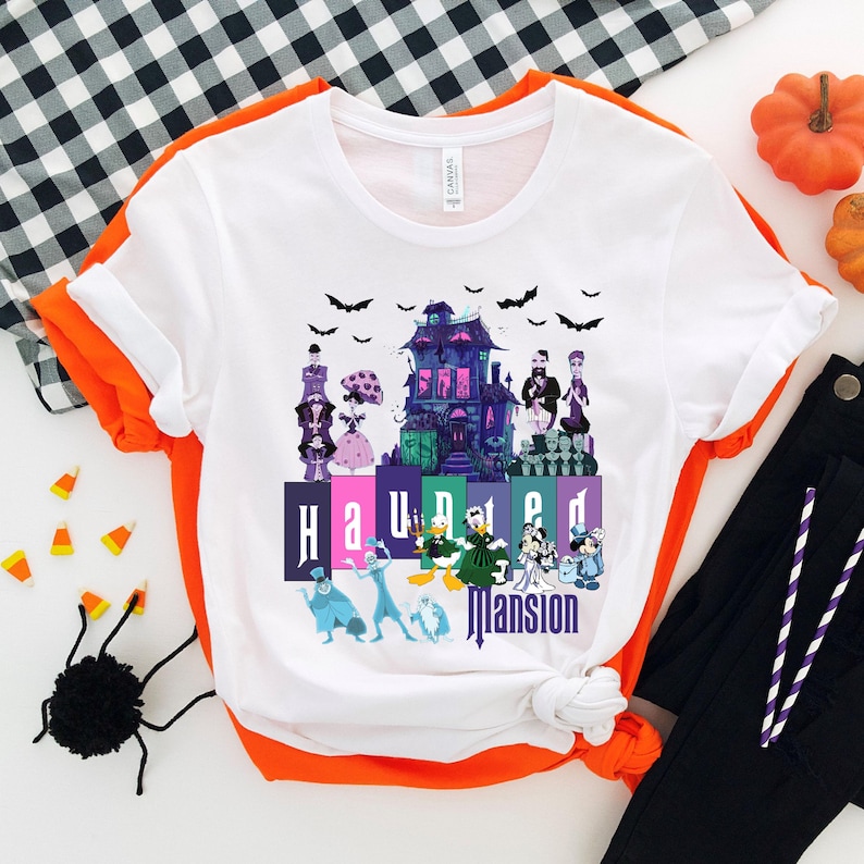 Puede incluir: Camiseta blanca con un gr&aacute;fico colorido de la Mansi&oacute;n Encantada de Disney. El gr&aacute;fico incluye el edificio de la Mansi&oacute;n Encantada, personajes del paseo y el texto "Haunted Mansion".