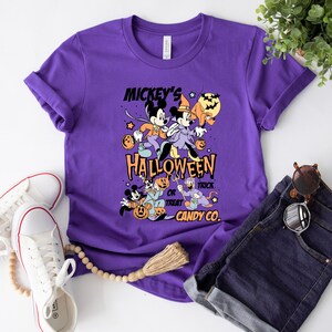 May include: Purple t-shirt with a vintage Mickey Mouse Halloween design. The design features Mickey Mouse, Minnie Mouse, Donald Duck, and Goofy dressed in Halloween costumes. The text on the shirt reads "Mickey's Halloween Trick or Treat Candy Co."