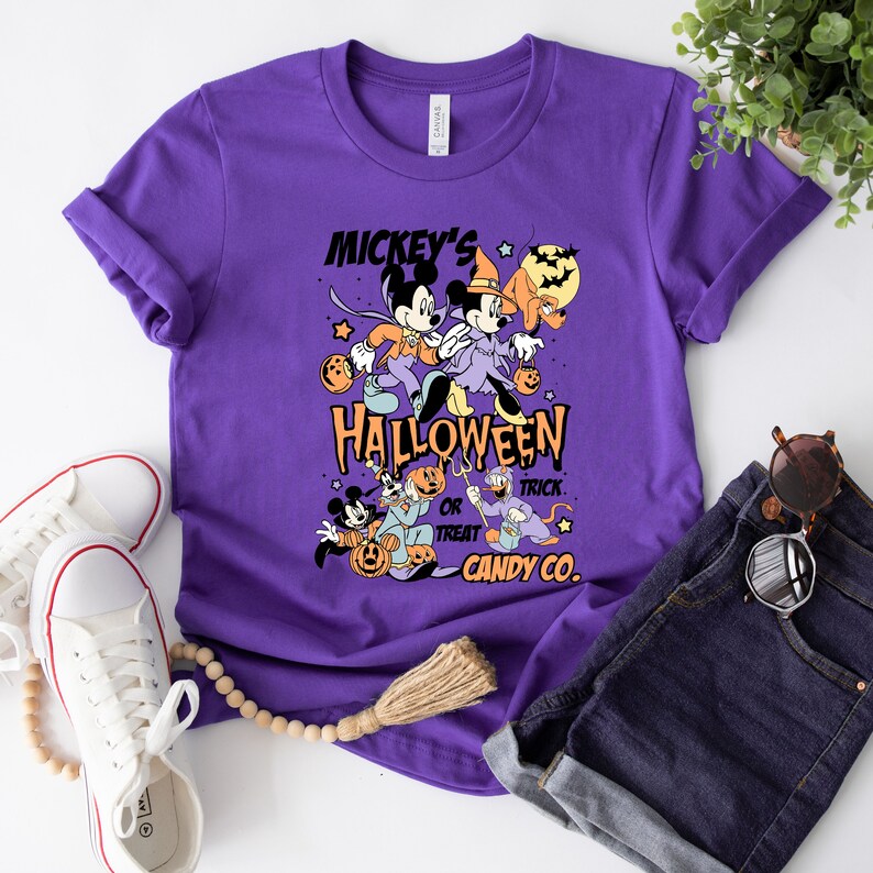 May include: Purple t-shirt with a vintage Mickey Mouse Halloween design. The design features Mickey Mouse, Minnie Mouse, Donald Duck, and Goofy dressed in Halloween costumes. The text on the shirt reads "Mickey's Halloween Trick or Treat Candy Co."