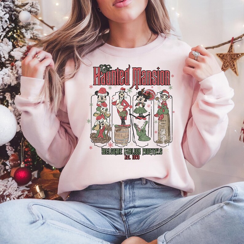 May include: Pink sweatshirt with a vintage-style graphic of the Haunted Mansion attraction at Disney World. The graphic features characters from the ride, including the Ghost Host, a hitchhiking ghost, and a singing bust. The text "Welcome Foolish Mortals Est. 1969" is printed below the graphic.