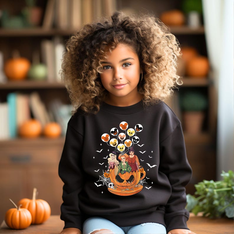 May include: A black sweatshirt with a graphic of three witches riding in a pumpkin-shaped cup with Mickey Mouse balloons. The witches are wearing purple, green, and blue dresses. The sweatshirt has a Halloween theme.