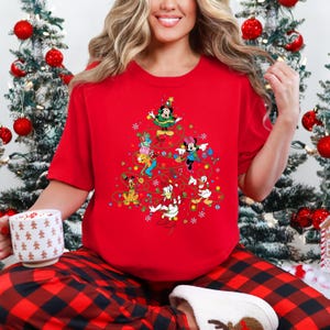 K&ouml;nnte beinhalten: Ein rotes T-Shirt mit einem Weihnachtsbaum-Design aus Disney-Figuren, darunter Mickey und Minnie Mouse, Donald Duck und Goofy. Das Shirt wird mit einer rot-schwarz karierten Pyjamahose kombiniert, was einen festlichen Look ergibt.