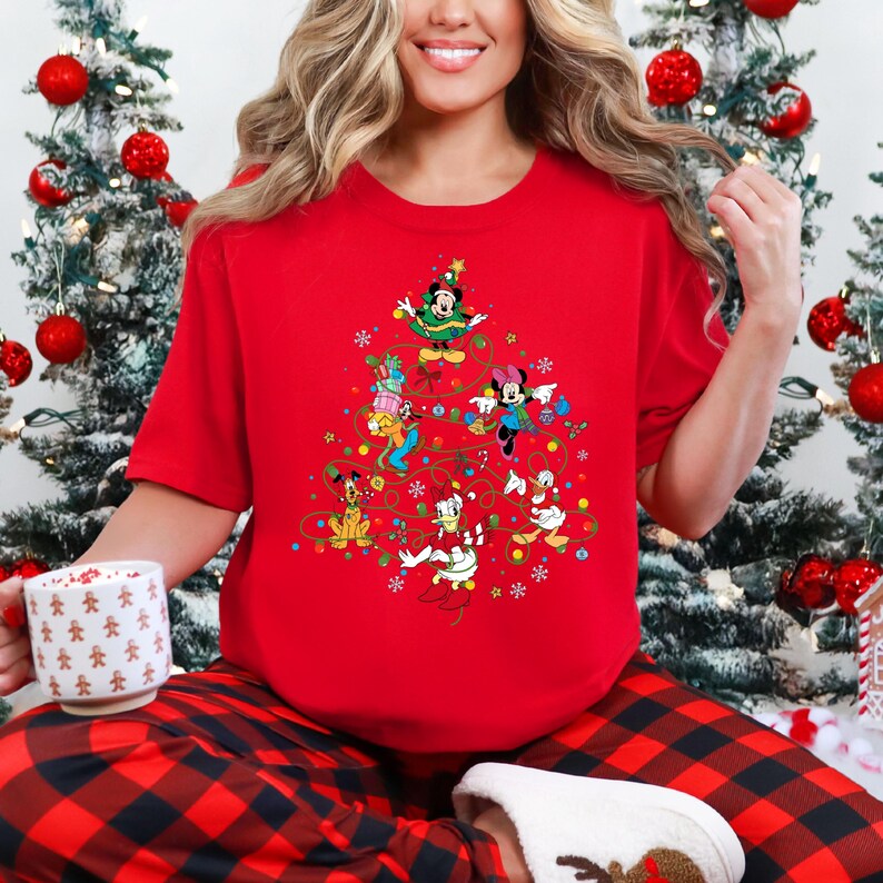 K&ouml;nnte beinhalten: Ein rotes T-Shirt mit einem Weihnachtsbaum-Design aus Disney-Figuren, darunter Mickey und Minnie Mouse, Donald Duck und Goofy. Das Shirt wird mit einer rot-schwarz karierten Pyjamahose kombiniert, was einen festlichen Look ergibt.