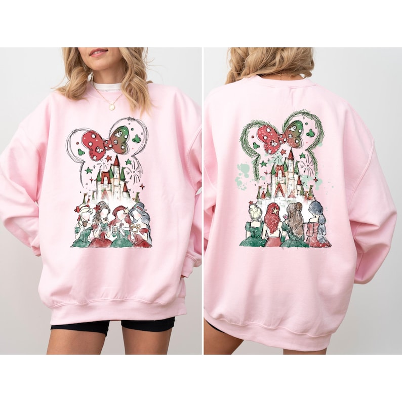 May include: A light pink sweatshirt featuring a whimsical design of a castle, fireworks, and several figures in dresses. The design includes a large bow and mouse ears. The back of the sweatshirt has a similar design.