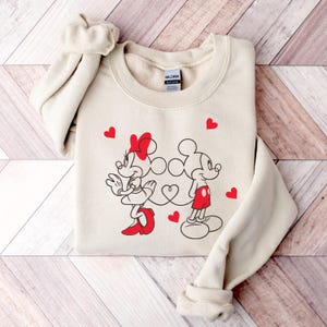 Op de afbeelding: Een beige sweatshirt met een afbeelding van Mickey Mouse en Minnie Mouse die hand in hand staan. De afbeelding is in zwart-wit met rode accenten. Er zijn rode hartjes rondom de afbeelding verspreid.