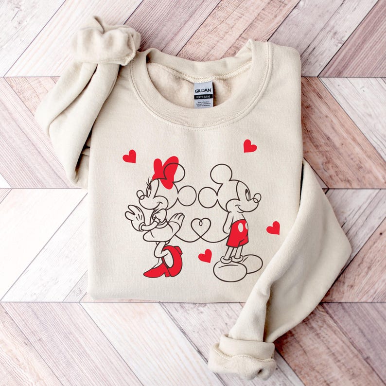 Op de afbeelding: Een beige sweatshirt met een afbeelding van Mickey Mouse en Minnie Mouse die hand in hand staan. De afbeelding is in zwart-wit met rode accenten. Er zijn rode hartjes rondom de afbeelding verspreid.