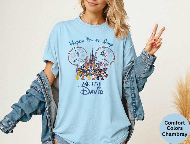 May include: Light blue t-shirt with a graphic featuring Mickey Mouse ears, fireworks, and a castle, with the text "Happy 4th of July" and "Est. 1776 David". The shirt is a Comfort Colors Chambray.