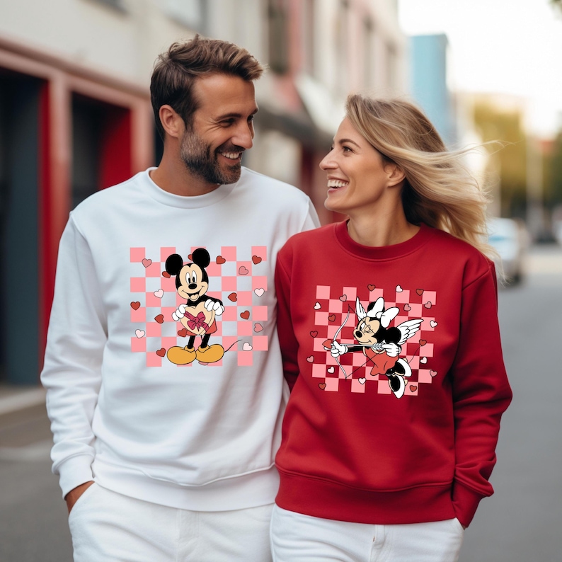 Pode incluir: Um casal usando su&eacute;teres combinando com tema do Dia dos Namorados. O su&eacute;ter do homem &eacute; branco com um padr&atilde;o xadrez rosa e branco e uma imagem de desenho animado do Mickey Mouse segurando uma caixa de chocolates em forma de cora&ccedil;&atilde;o. O su&eacute;ter da mulher &eacute; vermelho com um padr&atilde;o xadrez rosa e branco e uma imagem de desenho animado da Minnie Mouse com asas de anjo segurando um arco e flecha.
