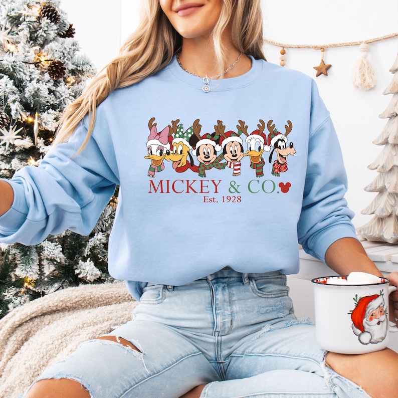 K&ouml;nnte beinhalten: Hellblaues Sweatshirt mit einem festlichen Design von Disney-Figuren mit Rentiergeweihen und Schals. Der Text "MICKEY & CO." und "Est. 1928" sind darunter gedruckt. Eine Tasse mit Weihnachts-Motiv wird gehalten.