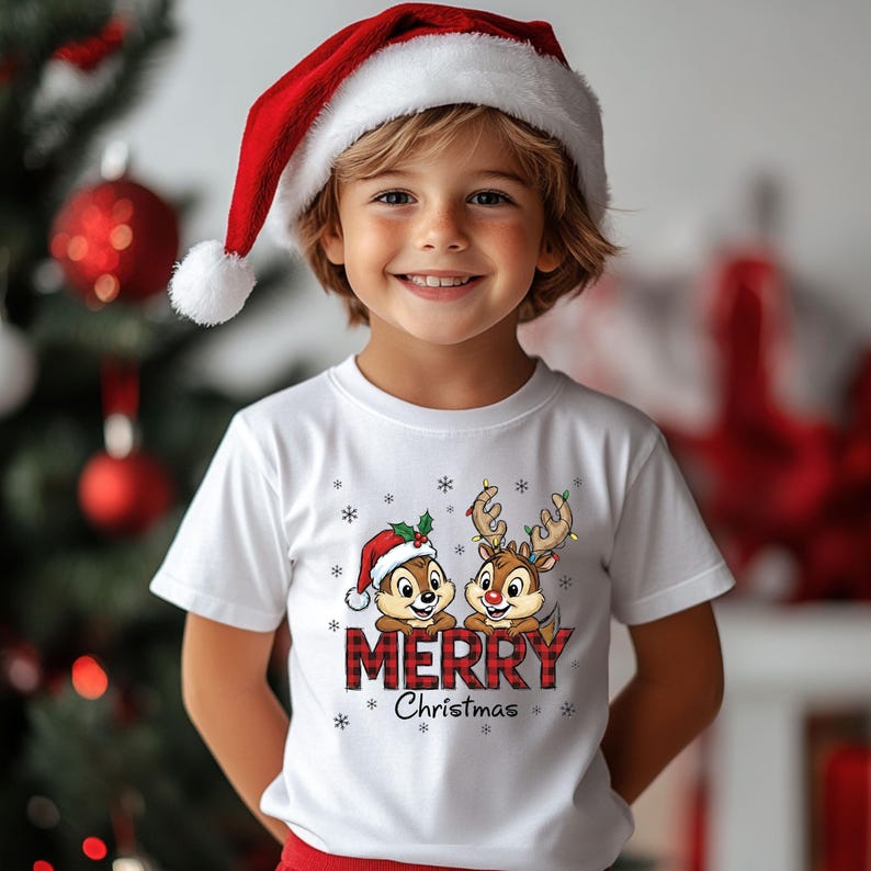 May include: A child in a white t-shirt with a festive Christmas design. The shirt displays two cartoon characters, one in a Santa hat and the other with reindeer antlers, and the words "Merry Christmas" in red plaid. The child is also wearing a red and white Santa hat.