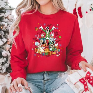 K&ouml;nnte beinhalten: Rotes Sweatshirt mit einem weihnachtlichen Disney-Motiv. Die Grafik zeigt Micky und Minnie Maus, Donald Duck und Goofy, umgeben von Geschenken, Schneeflocken und Unterschriften der Charaktere. Ein saisonales Kleidungsst&uuml;ck.