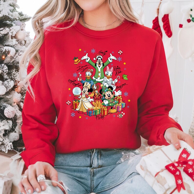 K&ouml;nnte beinhalten: Rotes Sweatshirt mit einem weihnachtlichen Disney-Motiv. Die Grafik zeigt Micky und Minnie Maus, Donald Duck und Goofy, umgeben von Geschenken, Schneeflocken und Unterschriften der Charaktere. Ein saisonales Kleidungsst&uuml;ck.