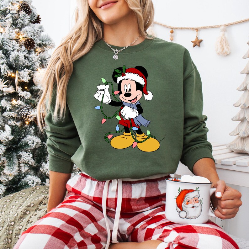 Puede incluir: Sudadera verde con un gr&aacute;fico de Mickey Mouse con un gorro de Pap&aacute; Noel y sosteniendo luces navide&ntilde;as. La sudadera tiene cuello redondo y mangas largas. Una taza blanca con un gr&aacute;fico de Pap&aacute; Noel se sostiene en la mano.