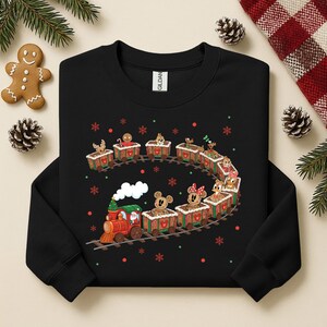 May include: Black sweatshirt featuring a festive train design. The train is composed of gingerbread-style cars, each carrying a different character. Red, green, and white accents add to the holiday theme. Snowflakes and dots surround the train.