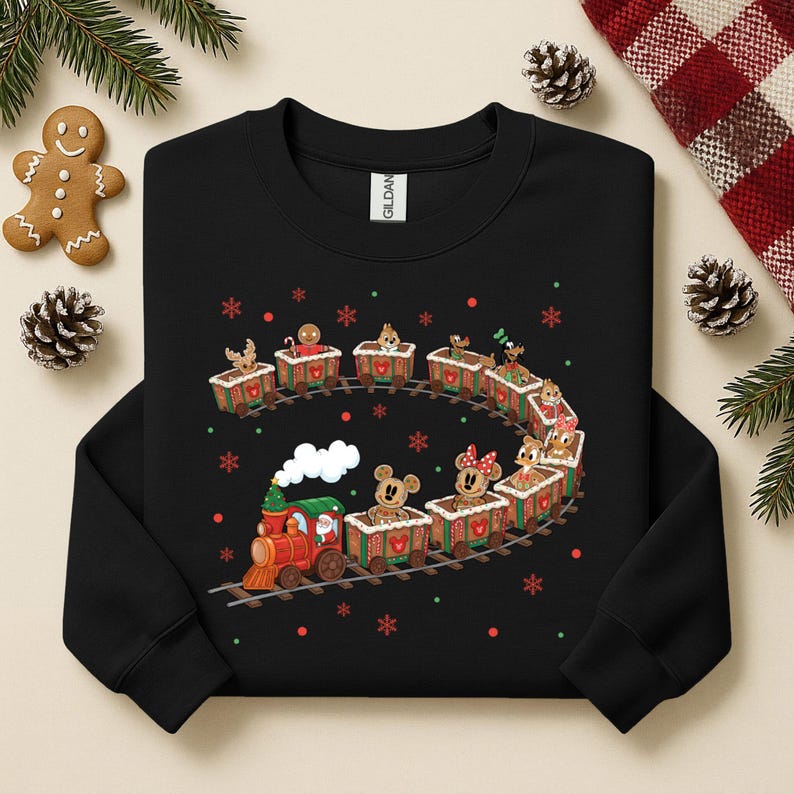 May include: Black sweatshirt featuring a festive train design. The train is composed of gingerbread-style cars, each carrying a different character. Red, green, and white accents add to the holiday theme. Snowflakes and dots surround the train.
