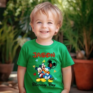 May include: A young child wearing a green t-shirt with the name "Jason" in red letters. The shirt features a graphic of Mickey Mouse, Donald Duck, and Goofy, with the number "3" and the words "Birthday Bop".