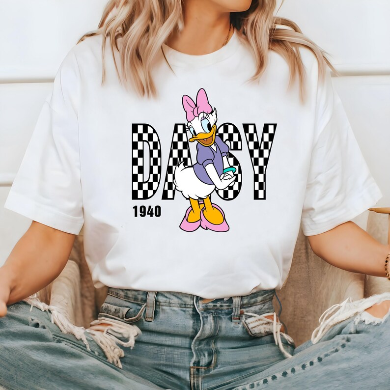 May include: White t-shirt with a black and white checkered design that spells out "DAISY" with a cartoon image of Daisy Duck in the center. The year "1940" is printed below the design.