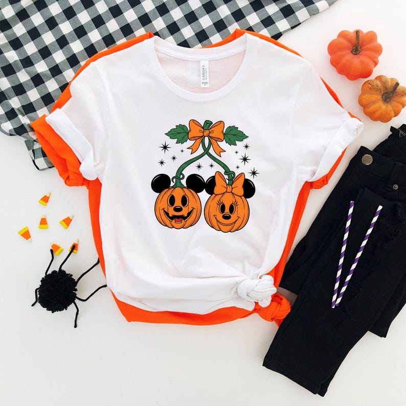 May include: White t-shirt with orange trim featuring a Halloween-themed design. The graphic shows two smiling jack-o'-lanterns with Mickey Mouse ears and bows, connected by green vines and an orange bow. Black pants and decorative pumpkins are also visible.