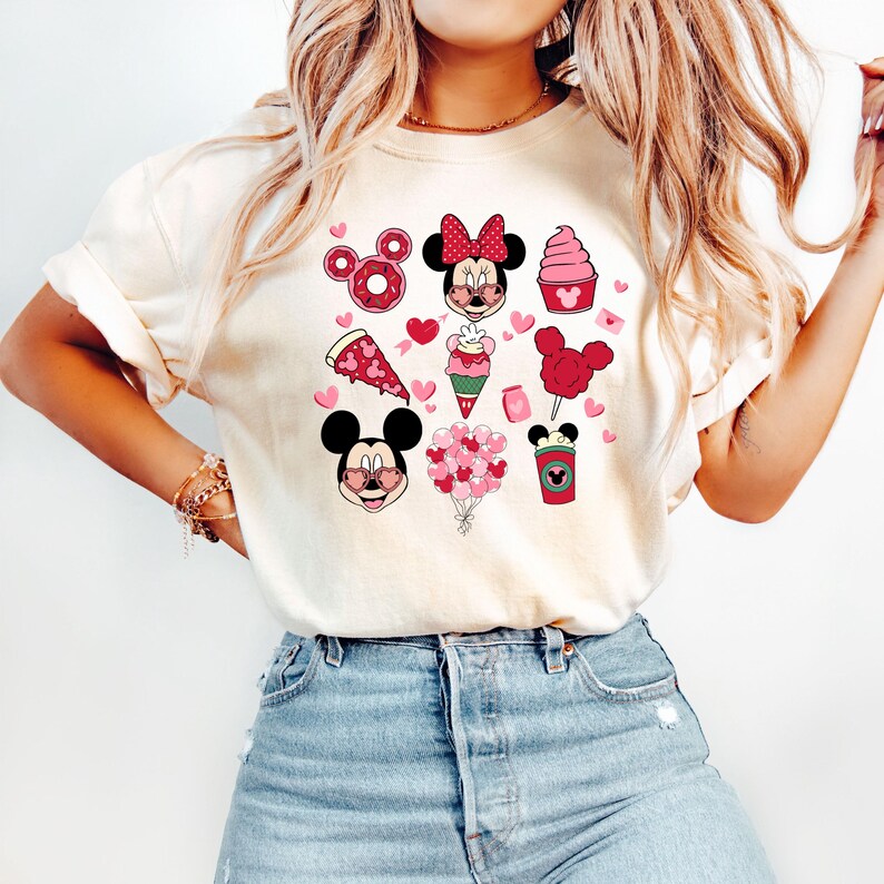 May include: A white t-shirt with a pink and red graphic design featuring Mickey Mouse and Minnie Mouse with hearts, balloons, and other Valentine's Day themed imagery.