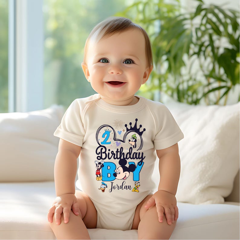 Op de afbeelding: Een wit rompertje met een blauw en zwart Mickey Mouse-ontwerp. Het ontwerp bevat het nummer 2, de woorden "Birthday Boy" en de naam "Jordan".
