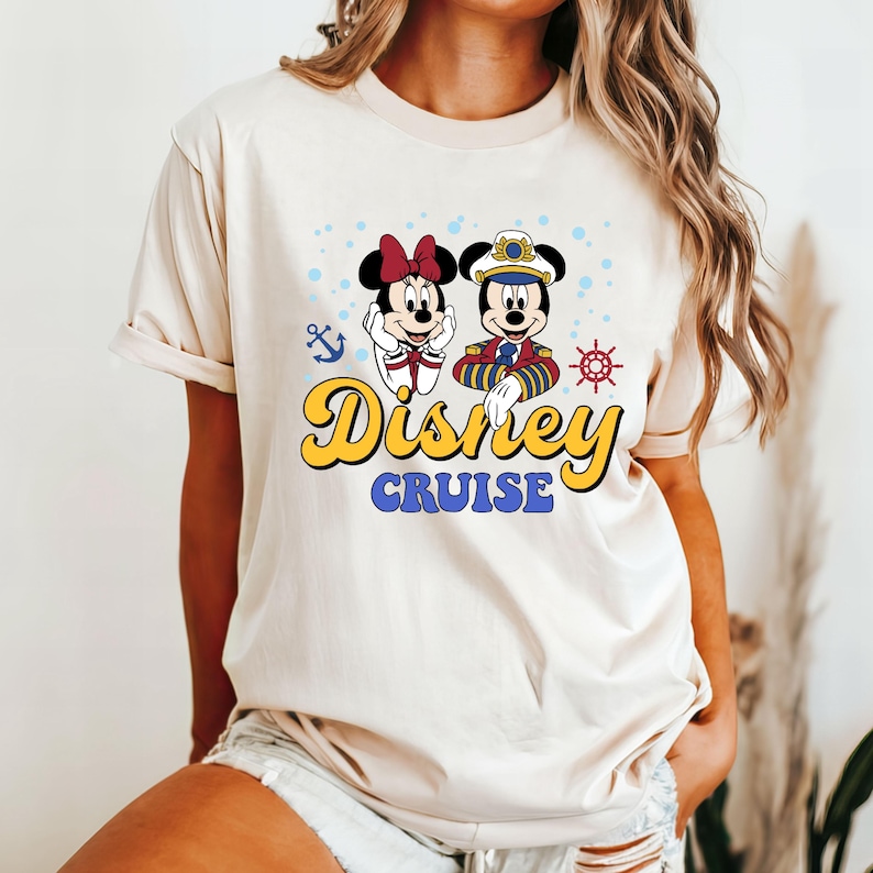 May include: A beige t-shirt with a graphic of Mickey and Minnie Mouse dressed as a captain and a sailor. The text "Disney Cruise" is printed in yellow below the characters.