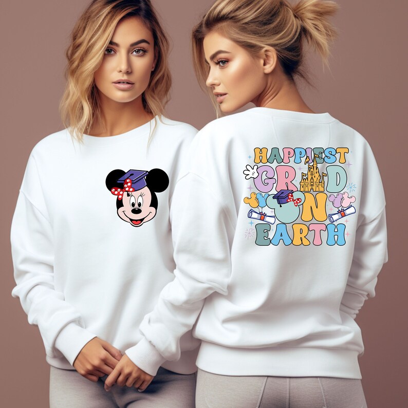 Puede incluir: Sudadera blanca con un gr&aacute;fico colorido en la espalda que dice "Happiest Grad on Earth" con un gorro de graduaci&oacute;n, orejas de Mickey Mouse y un castillo. La parte delantera de la sudadera tiene un personaje de Mickey Mouse con un gorro de graduaci&oacute;n.
