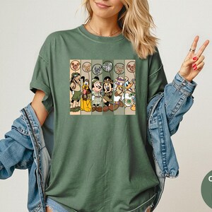 May include: Moss green t-shirt featuring a graphic of Disney characters in safari outfits. The graphic includes Goofy, Pluto, Mickey Mouse, Minnie Mouse, Donald Duck, and Daisy Duck. The shirt has a relaxed fit and short sleeves. The text "Comfort Colors Moss" is on the bottom right.
