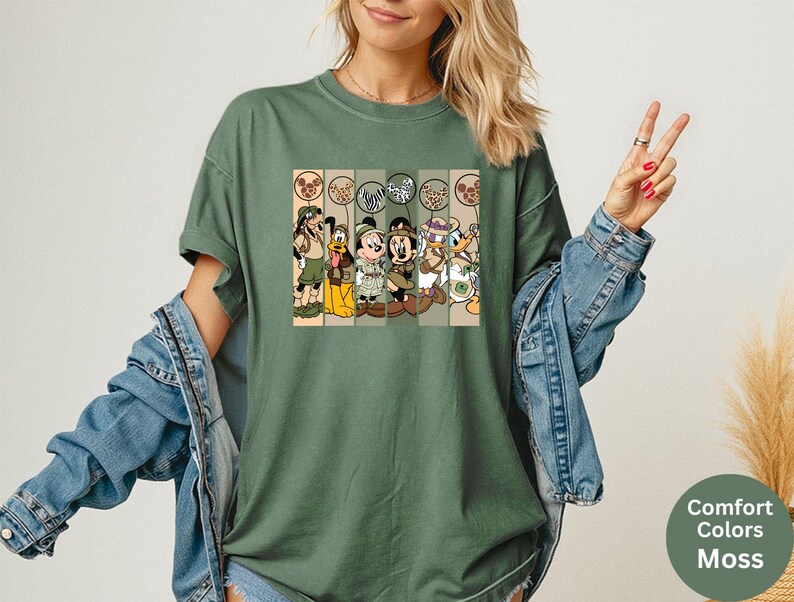 May include: Moss green t-shirt featuring a graphic of Disney characters in safari outfits. The graphic includes Goofy, Pluto, Mickey Mouse, Minnie Mouse, Donald Duck, and Daisy Duck. The shirt has a relaxed fit and short sleeves. The text "Comfort Colors Moss" is on the bottom right.