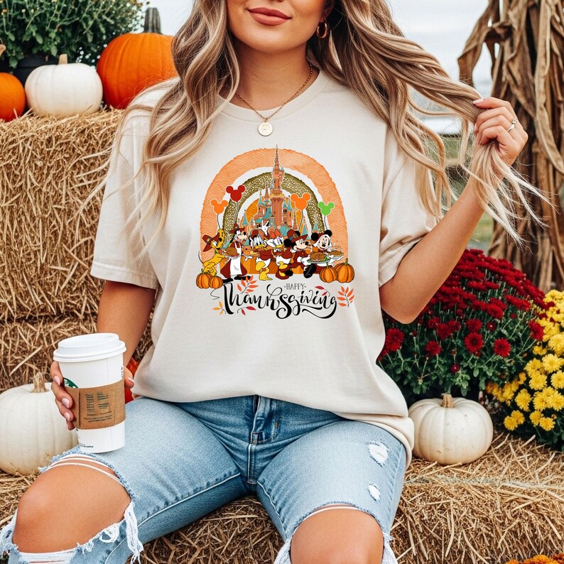 May include: Beige t-shirt featuring a colorful graphic of Disney characters, a castle, and pumpkins, with the text "Happy Thanksgiving". The design is set against a rainbow arch. The shirt is worn by a person.