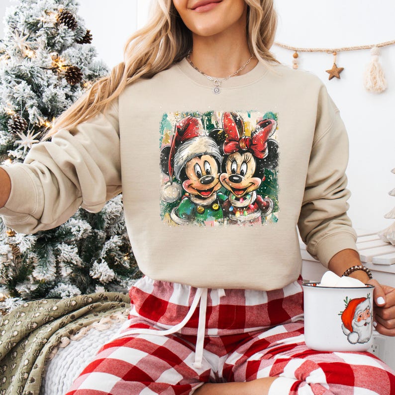 May include: Beige sweatshirt featuring a colorful graphic of Mickey and Minnie Mouse in Christmas attire. Mickey wears a Santa hat and green coat, while Minnie dons a red bow and dress. The sweatshirt is paired with red and white plaid pajama pants.