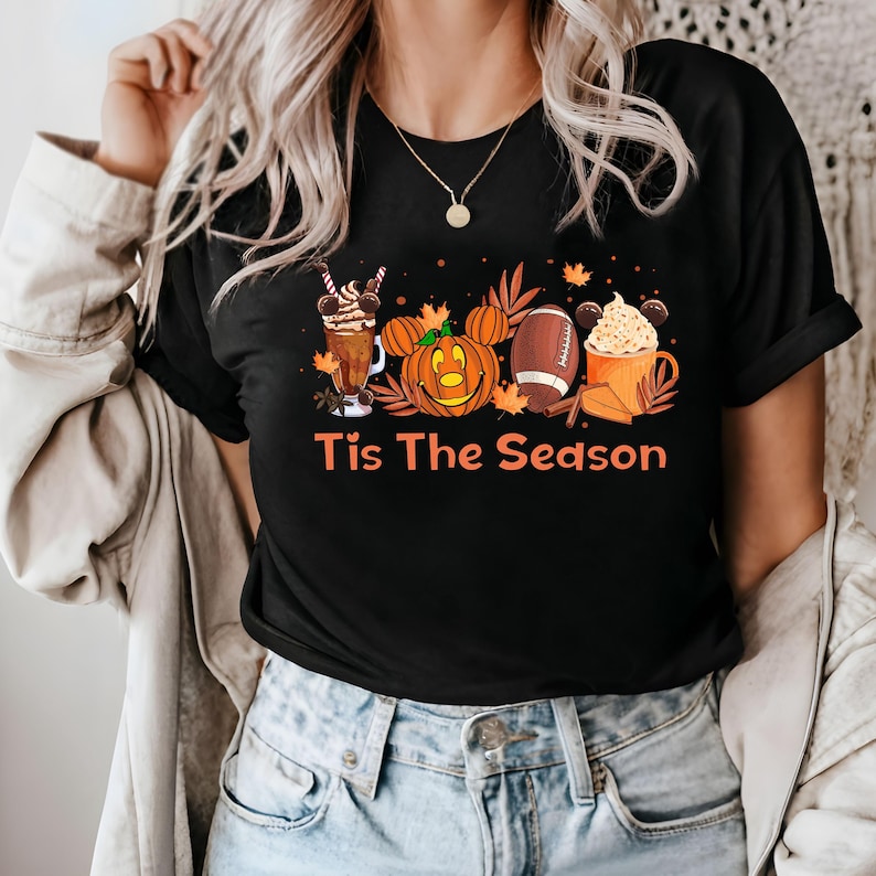 K&ouml;nnte beinhalten: Schwarzes T-Shirt mit einem Aufdruck einer K&uuml;rbis mit Mickey-Maus-Ohren, einem Football, einem Milchshake und einem hei&szlig;en Kakao. Der Text "Tis The Season" ist unter dem Aufdruck gedruckt.