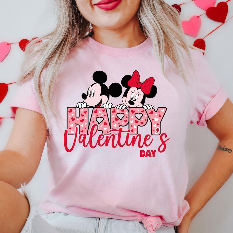 May include: Pink t-shirt featuring a graphic of Mickey Mouse and Minnie Mouse peeking over a heart-shaped banner that reads "Happy Valentine's Day".