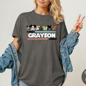 May include: Dark gray t-shirt featuring a graphic design with cartoon character heads, lightsabers, and the name "GRAYSON" in white. The design includes a red and a blue lightsaber. The shirt is a Comfort Colors Pepper tee.