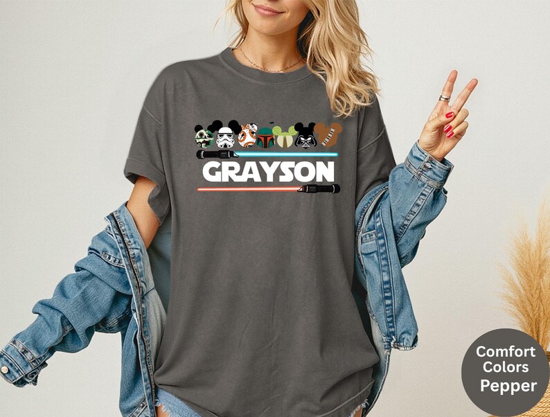 May include: Dark gray t-shirt featuring a graphic design with cartoon character heads, lightsabers, and the name "GRAYSON" in white. The design includes a red and a blue lightsaber. The shirt is a Comfort Colors Pepper tee.
