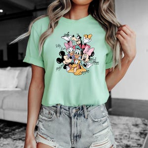 May include: A light green t-shirt featuring a colorful graphic of Disney characters including Mickey Mouse, Minnie Mouse, Donald Duck, Daisy Duck, Goofy, and Pluto. The characters' names are written in a script font.