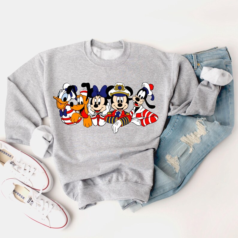 Peut inclure: Un sweat-shirt gris avec un imprim&eacute; graphique de Mickey Mouse, Minnie Mouse, Donald Duck, Dingo et Pluto habill&eacute;s en marins. Les personnages sont tous souriants et ont des accents rouge, blanc et bleu.