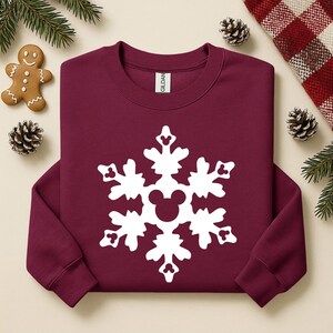 May include: Burgundy crewneck sweatshirt with a large white snowflake design. The snowflake features a Mickey Mouse head in the center. The sweatshirt is a festive holiday item, perfect for winter wear.